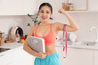 © Pixel-Shot - Sporty young Asian woman with tape measure and weight scales showing muscles in kitchen. Diet concept