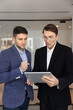 © fizkes - Two businessmen in suits standing indoor, analyze or discuss important data on digital tablet, focused on business strategy planning or problem-solving in corporate office. Modern tech, AI tools usage