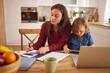 © Stockphotodirectors - Mother focuses on work using papers and pen at the kitchen table while child engages with a tablet nearby. Fresh fruits sit in a bowl, creating a warm home atmosphere.