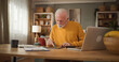 © Stockphotodirectors - An older man sits at a wooden desk in his living room. He focuses on filling out life insurance paperwork while using a tablet and has a laptop nearby. A banana sits on the table.
