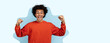 © Prostock-studio - A young individual with curly hair stands in front of a light blue background. They raise their fists in the air while smiling widely. The scene conveys a strong sense of joy.
