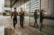 © qunica.com - Two professionals walk beside a glass-fronted building, reviewing documents and tablet. A business pair chats as they move along an urban sidewalk with reflections in the windows.