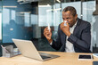 © Liubomir - Businessman sneezing into a tissue, holding a nasal spray bottle, experiencing allergy or cold symptoms at his office desk, indicating sickness impacting productivity