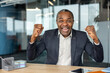 © Liubomir - Happy mature businessman sitting at a desk in a modern office, raising his fists in a gesture of success and excitement, celebrating a professional victory or great news
