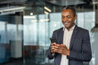 © Liubomir - Executive man standing in a modern office building, happily using his mobile phone for professional communication and networking, representing success and technology in the contemporary workplace