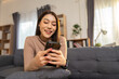 © Supachai - Happy Asian woman freelancer using smartphone sitting on sofa at home. Smiling female relaxing in living room browsing social media or chatting online concept