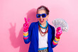 © deagreez - Energetic mature glamour woman in vivid blue jacket pink gloves and oversized blue glasses flashes peace sign while holding cash in a fun retro vibe photoshoot