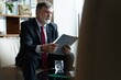 © opolja - Mature businessman in formal suit concentrating on reading the report, sitting in the lobby and preparing for the meeting.
