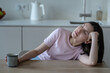 © ingoodtime - Tired young woman sitting at table with cup of coffee, leaning on hand with eyes closed. Morning fatigue, low energy, exhaustion. Sleepy female rest head on hand while holding mug, feeling drained.