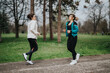 © qunica.com - Two women jog together in a park, wearing jackets and athletic leggings. They move along a path in a green, leafy setting, enjoying a casual outdoor workout.