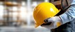 © Mariia - Worker holding bright yellow safety helmet in hands against blurred construction site background. Horizontal banner with copy space for text, building industry and workplace safety concept