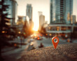 © andyaziz6 - Urban city street scene at sunset with a vibrant orange location pin marker on a textured concrete surface, featuring modern glass and steel buildings in the background, warm