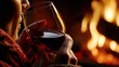 © Giovanna - Close view of a woman holding a glass of red wine by a glowing fire, enjoying a warm cozy evening at home. Soft light and rich tones create a romantic winter mood of comfort and relaxation.