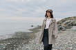 © SHOTPRIME STUDIO - Woman in stylish coat and hat walks on rocky beach under cloudy sky. Concept of autumn fashion, casual outfit, and outdoor lifestyle by the sea with natural mood and calm atmosphere.
