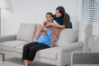 © Thitaporn - An Asian mother and daughter sit closely together on a sofa at home, sharing a tender embrace and warm smiles, expressing love, gratitude, and deep family connection in a bright living room
