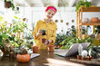 © sofiko14 - A woman smiles while working on her plants, surrounded by greenery and using a laptop