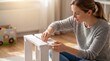 © JONATHAN - Focused young woman assembling white wooden furniture at home, carefully attaching parts of a small stool or shelf. Engaging in a DIY home improvement project in soft natural light.