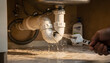 © Oleksandr - Close-up of a broken and leaking sink trap, with water spraying out as a person attempts to fix it with an adjustable wrench under a kitchen counter