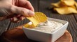 © IkmotIQ - Crispy potato chip dipping into creamy white sauce, hand holding salty snack over bowl of garlic dip, crunchy appetizer on rustic wooden table