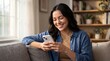 © Muhammad - Smiling young woman with dark hair wearing a denim shirt, happily using her white smartphone while relaxing on a gray sofa in a brightly lit living room.