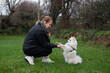 © Мар'ян Філь - A woman crouches on green grass teaching her white dog to give a paw, practicing obedience and bonding during a chilly overcast walk in park. Woman training her white dog outdoors