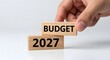 © saba - Hand carefully placing wooden block labeled budget 2027 on top of another block with year displayed.