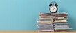 © oksa_studio - Alarm clock sitting on a high stack of documents and files, representing office workload, deadlines, stress, and time management against a blue background
