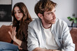 © luciano - Young couple sitting back to back on couch having a dispute, expressing relationship conflict, unresolved problem or question, emotional distance, or communication problems in a modern living room.
