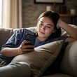 © Ali - Young woman relaxing on a comfortable sofa at home, engrossed in her smartphone with a soft glow illuminating her face in a cozy living room setting.