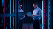 © Gede - Two IT professionals working with a laptop in a data center with neon lighting and server racks.