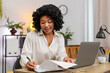 © Andrii Iemelianenko - African American businesswoman at home office matches documents to laptop data smiling satisfied. Freelancer girl at table ticks paper list with pen very pleased by accurate results feeling organized