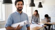 © Muhammad - Smiling bearded male architect holding rolled blueprints in a bright office, with colleagues collaborating on designs in the background. The man wears a light blue button-up shirt and looks
