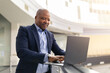 © Anastasiya - Focused mature black businessman working on laptop near office railing, modern interior with natural light and productive executive atmosphere
