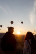 © Aleksei Toropov/Stocksy - A young couple stands against a backdrop of rising hot air balloons.
