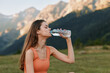 © SHOTPRIME STUDIO - A young woman drinks water from a bottle in a scenic mountain landscape. She wears a sleeveless top, with sunlight highlighting the scene and a calm, refreshing atmosphere.