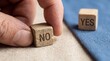 © Teresa - A hands fingers move a wooden block displaying NO on a textured light fabric Another YES block is visible in the blurred background on a blue textile surface