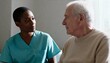 © Anna - A compassionate Black nurse listens to an elderly male patient during a consultation. A healthcare professional provides support and care in a nursing home or hospital setting.