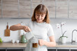 © Pixel-Shot - Young woman pouring milk from jug into glass with iced coffee in kitchen