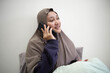 © airdone - Young Asian woman in hijab sitting on bed and talking on smartphone, smiling while having a phone call, cozy indoor lifestyle concept