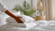 © dinatychynska - Person arranging fresh white pillow on clean bed with soft white bedding in bright minimalist bedroom with green plant, perfect hotel housekeeping and home comfort concept.