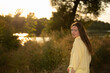 © Oleksandr Zinchenko - A woman is standing by a body of water at sunset, wearing a yellow shirt. She looks back with trees and grass around her