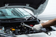 © Kannadhee - Close-up of a professional automotive mechanic holding a wrench and inspecting a car engine under the open hood. Concept of vehicle maintenance, car repair service, mechanical inspection, and automoti