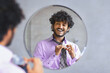 © Prostock-studio - A man looks at his reflection in a round mirror while he adjusts his tie. He wears a light purple shirt and has curly hair. The setting appears tidy and well-lit with a simple background.