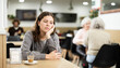 © JackF - In middle of cafe hall, sad girl is sitting at table and drinking coffee. Visitor to food court is sitting at table, eating light breakfast, coffee break