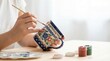 © Stitch - Artist's hands painting a traditional blue and red floral pattern on a ceramic mug.