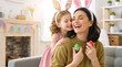 © Milan - Cheerful mother and daughter with Easter eggs wearing  bunny ears. Happy mom and child celebrating holiday. Mother holds colorful painted Easter eggs while her daughter hugs her from behind