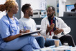 © DC Studio - Black male doctor in lab coat speaks with patient during clinic appointment, reviewing documents and discussing health concerns. Female nurse takes notes as physician explains treatment information.