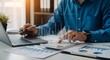 © Msthosneara - A professional businessman works diligently on his laptop at an office desk, surrounded by financial accounting documents and reports while writing notes in a notebook