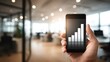 © OngCaLucK - Hand holding smartphone displaying growth chart against modern office background, symbolizing business success and technological advancement in analytics