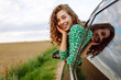 © maxbelchenko - Cheerful woman leans out window, enjoying rural landscape on sunny day. Young traveler in bright dress and hat feels free, catching wind in hair from car window. Concept: transportation, adventure.
